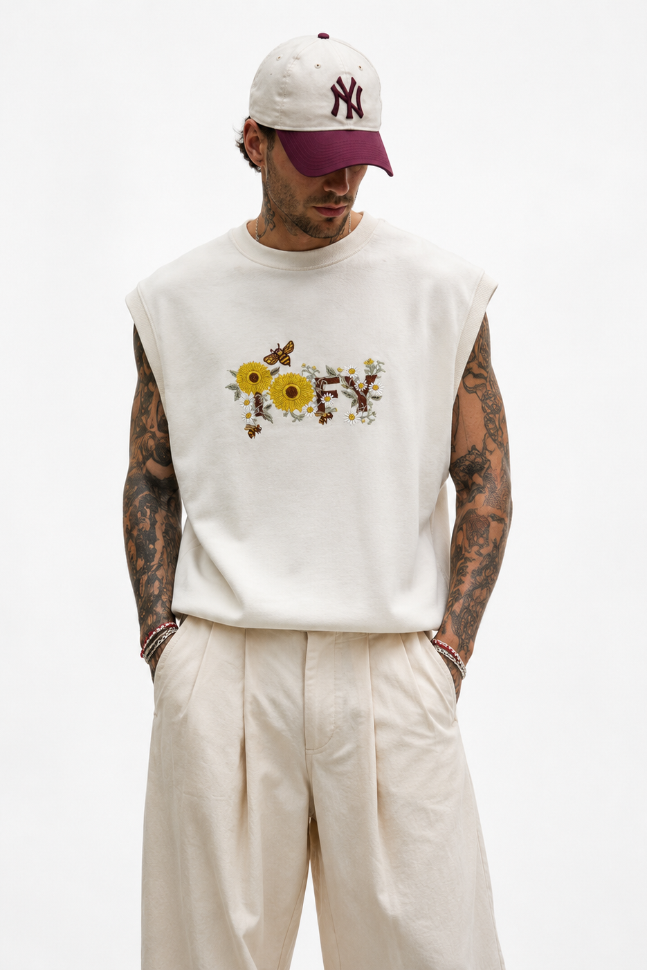 A man wearing a sleeveless white t-shirt with a yellow and brown floral graphic on the chest, paired with cream pleated trousers and a white baseball cap with a burgundy brim.