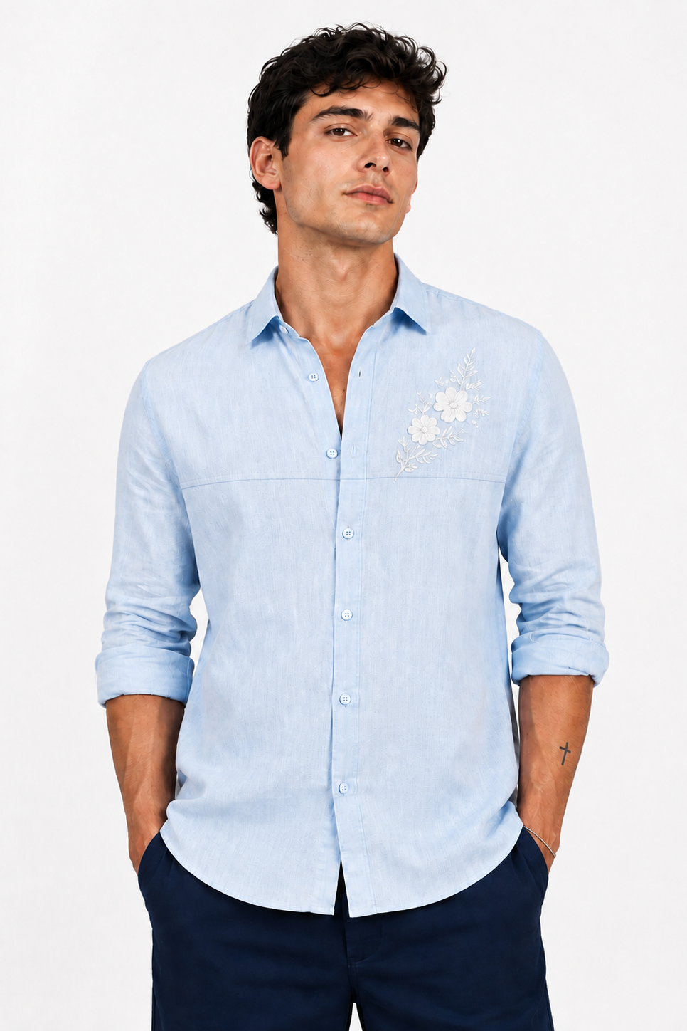 A man wearing a light blue linen button-down shirt with white floral embroidery on the left chest, paired with navy blue trousers, against a plain white background.