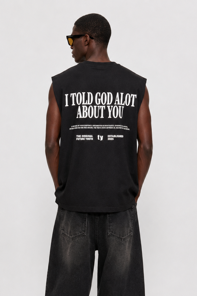 A man viewed from the back wearing a black sleeveless t-shirt with bold white text on the back, paired with baggy black jeans and black-and-white sneakers, against a light gray studio background.