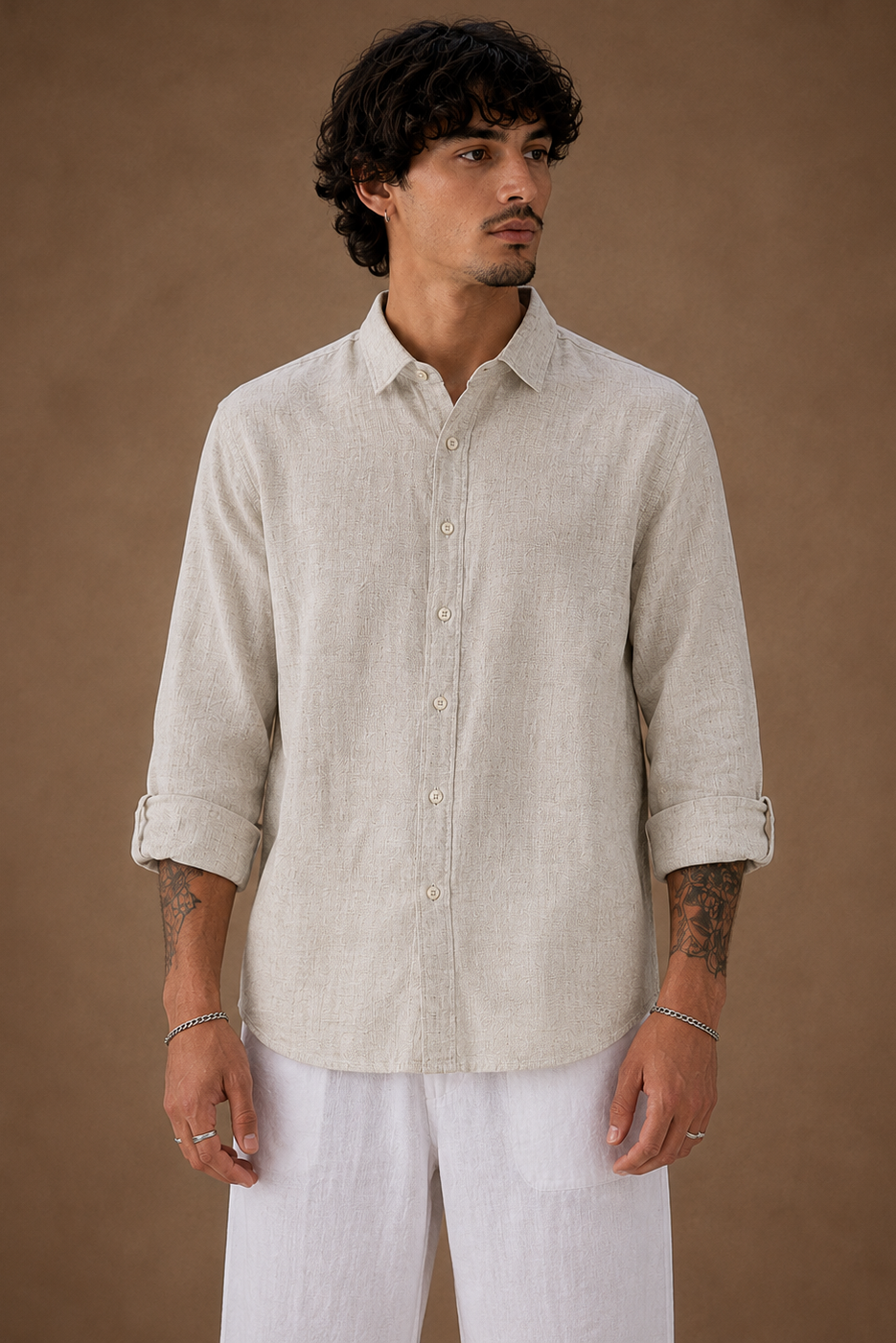 A man wearing a light beige linen button-up shirt with rolled-up sleeves and white pants, standing against a solid brown studio background.